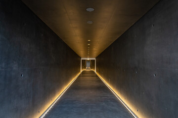 A long dark underground corridor made of artistic concrete stretching into the distance with lighting in the corners with a metal door at the end.
