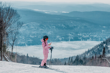 Phone on ski vacation - Woman skier using phone app on ski trail slope in amazing winter nature landscape. Girl looking at mobile smartphone wearing awesome ski clothing, helmet and goggles - obrazy, fototapety, plakaty