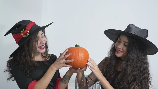 Gorgeous Girlfriends In Halloween Witch Hat Costumes Holding And Feeling Smooth Texture Of An Orange Pumpkin. medium shot