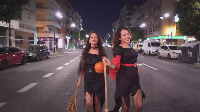 Girls In Witch Costumes Walking At The Road And Holding Pumpkin And Broomsticks. wide shot