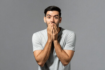 Shocked surprising young Caucasian man with hands covering mouth in isolated light gray studio...