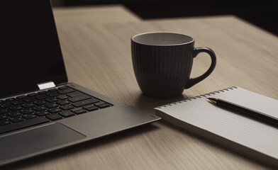 Close-up pencil on a notebook next to a laptop and cup of coffee on a beige wood table. Horizontal view.