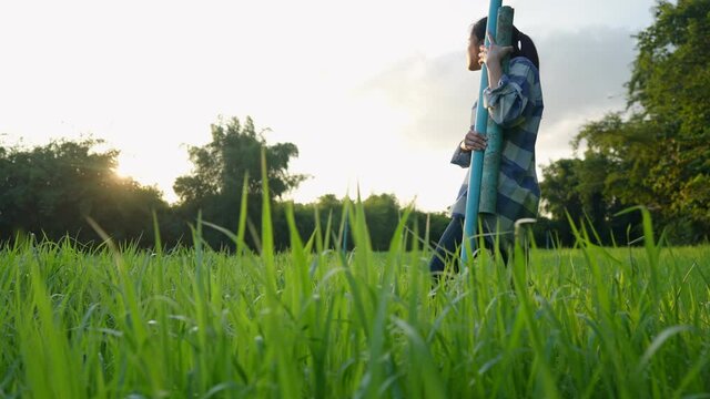 Young asian country girl holding blue pvc pipe going on the crop grass field get ready for work inside organic farm , sustainable agriculture, diy working equipment, simple domestic asian life