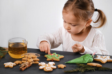 Child cooking and eating home made gingerbread cookies, stars, man. Happy toddler girl celebrated Christmas eve at home, Kid decorating pastry with icing. Copy space over plain wall