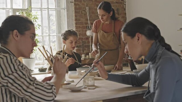 Slowmo handheld shot of young diverse people sitting at wooden table in groups chatting while painting ceramic items during pottery masterclass