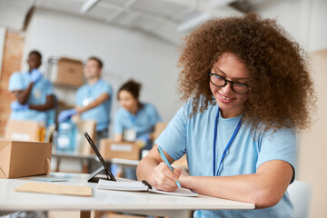 Portrait of young female volunteer in blue uniform making notes while working on donation project. Team sorting, packing items in the background
