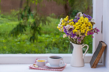 Old book, colorful bouquet of flowers and white cup of tea on background of window with raindrops at home at summer day