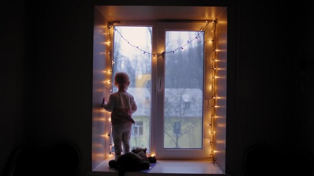 Child Looking out Window During Snow. A little baby at Christmas night. Girl looking through the window. A child stands at the window against the background of Christmas decorations. A lonely child.