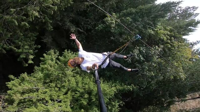 Vertical action camera shot of a young man, happily swinging out from the platform up in the trees, strapped to a zipline and lying down and stretching his arms and legs out in freedom