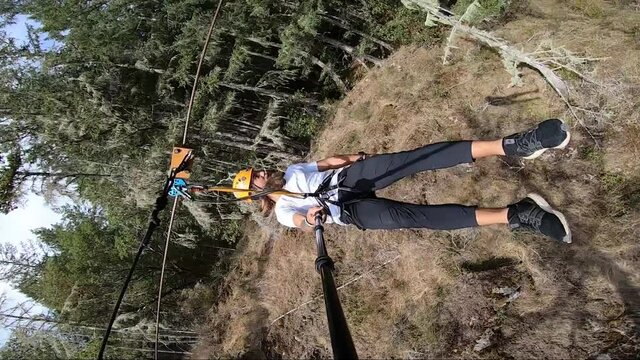 Vertical action camera shot of a young man swinging by very close to trees and bushes in the Canadian forest on the zipline in the hard summer sun on his holiday. He smiles during the thrilling ride.
