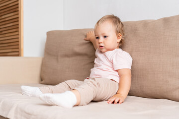 Fototapeta premium Little girl sitting on sofa and looking away with interest. Blond blue-eyed baby in light clothes