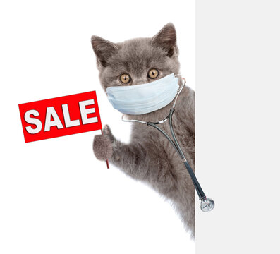 Kitten wearing stethoscope and medical protective fsce mask looks from behind empty banner and shows sales symbol. Isolated on white background