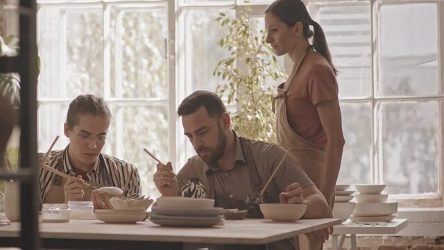 Slowmo shot of young female potter helping two men sitting at wooden table during ceramics masterclass in cozy workshop