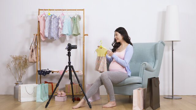 asian pregnant influencer sitting on a chair is talking and feeling the textile while making a video about baby clothes at white background.
