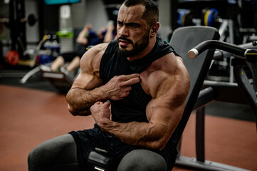 strong bearded caucasian man bodybuilder watching his shoulder and arm muscle sitting in sport...