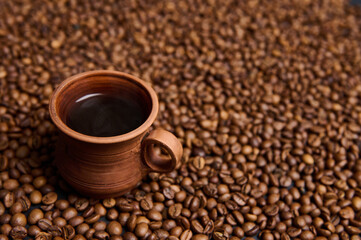 Close-up composition of a clay coffee cup with freshly brewed coffee drink with steam on the background of freshly roasted coffee beans with copy space for advertisement. Still life