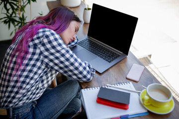 Young woman lying on her arms on the table in cafe in front of laptop with cup of coffee, sleepy.