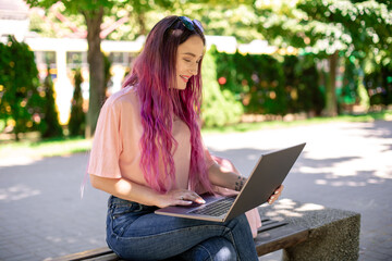 Young girl is studying in the spring park, sitting on the wooden bench and browsing on her laptop