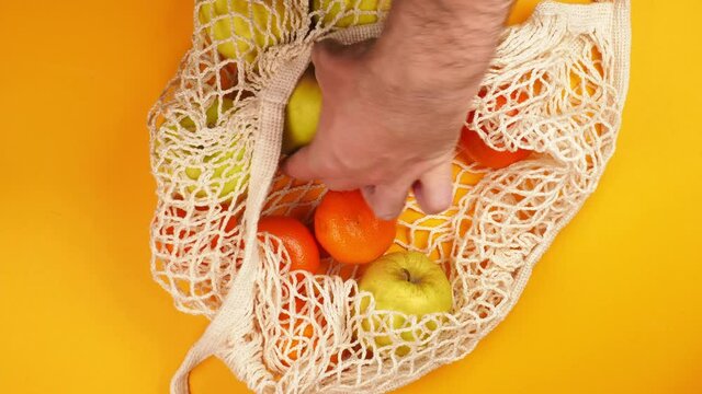 Male hand picks ripe juicy orange tangerines and green apples from an eco string bag on a bright yellow background