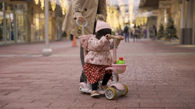 Lovely Japanese Baby Girl Having Fun Riding Tricycle With Her Mom’s Help On A Snowy Pedestrian Street With Holiday Lights And Christmas Decorations On Background