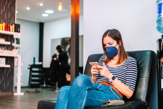 Woman Sitting In Waiting Room Using Phone And Wearing Surgical Mask At Beauty Salon