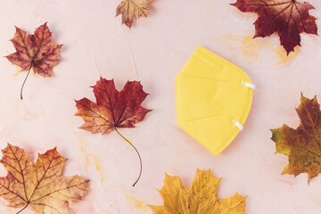 Yellow respirator KN95 or FFP2 mask on pink background, autumn maple leaves, copy space. Fall and...