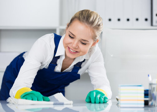 Young woman cleaner in overall wiping desk in modern office