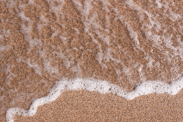 Background of a sandy beach with a sea wave. Foam from a wave on the sand. A foamy ocean wave on a seashell beach