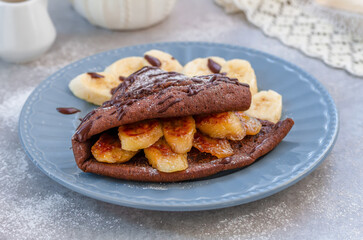 Chocolate dutch baby served on a plate with sliced caramelized bananas and melted chocolate. Close up, selective focus, gray concrete background.