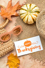 Wicker bag, hat, dry leaves, pumpkin, sunglasses and paper card with text HAPPY THANKSGIVING on color background
