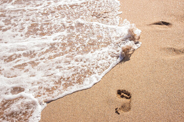 Human footprints on the sand with sea water. Foam from a wave on the sand. A foaming ocean wave washes away the traces on the shell beach