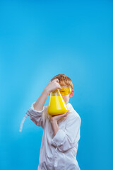 Schoolboy wearing protective eye glasses and gown holding chemical container on blue background