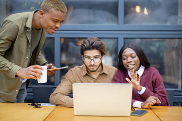 Multiracial students having video call on laptop at table outdoors. Concept of remote and e-learning. Idea of students lifestyle. Smiling young indian man, black girl and guy. University campus