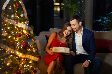Smiling young couple sitting together on couch and sharing with presents. Young woman wearing red...