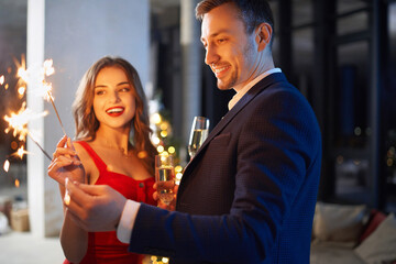 Handsome man in suit and beautiful woman in red dress holding sparklers and glass of champagne. Young couple enjoying christmas evening together at home.