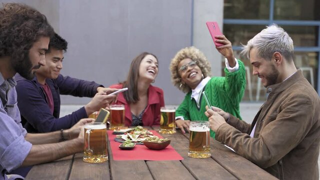 Group of happy multi ethnic friends with smartphones taking picture of food and drink, and selfie at outdoors bar to share it on social media apps
