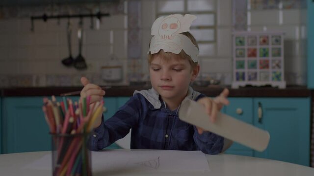 Kid playing pirate's game with paper telescope and headwear with skull and crossbones. Preschool boy drawing with color pencils sitting alone at home.