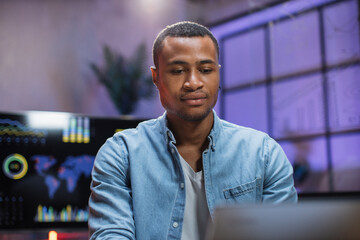 African american man in denim shirt sitting at office desk and working on wireless laptop. Handsome guy using modern gadgets at work. Business and finance concept.