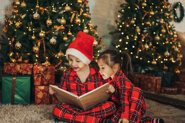 Children in matching pajamas are reading book on the floor in front of the Christmas tree
