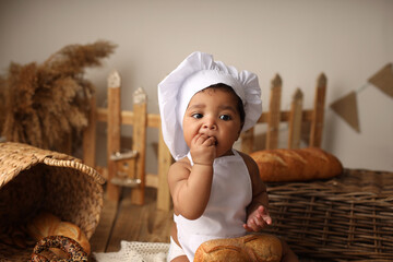 a cute dark-skinned kid with curly hair in a chef's costume has a bagel and a roll. High-quality photography