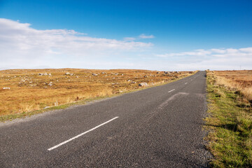 Fototapeta na wymiar Small narrow asphalt road in a huge empty fields. Clean blue cloudy sky. Warm sunny day. Connemara, Ireland. Travel and transportation concept