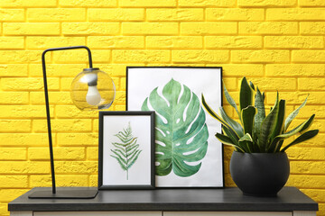 Beautiful plant in pot, lamp and pictures on black table near yellow brick wall. Interior accessories
