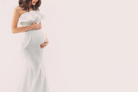 Happy pregnant woman in white dress gently hugs touch her belly with hands. Light beige studio background. Beautiful pregnancy, motherhood, expecting a baby, family health concept. Free space for text
