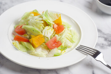 Salad with cabbage, tomatoes, cucumber, pumpkin in a plate on the marble table. Behind there is a gravy boat with sauce. Breakfast