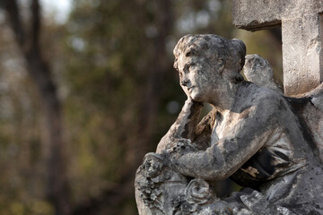 A sitting, pensive Angel. An angel with his head on his hand, looking at the sky. Blurred trees in...