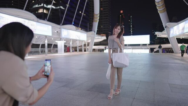 A two cheerful asian girl friends having fun together taking photos of her friend with modern beautiful concrete city decorating bridge, hanging out on weekend, shopping time, femininity society