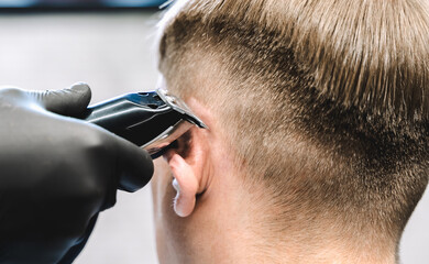 Visit to the barbershop. Hairdresser, barber shaves client's temples,sides with hair clipper. Young stylish man makes fashionable haircut.Customer sitting in dark men's beauty salon covered with cape