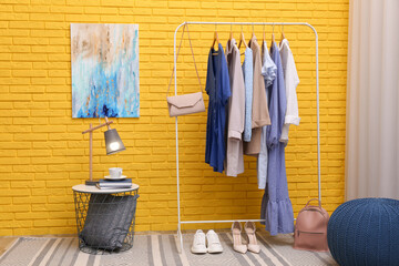 Rack with different stylish clothes and shoes near yellow brick wall in room