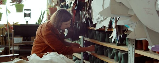 Caucasian woman choosing and taking shoe last from shelf in shoemaking workshop