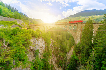 Fototapeta na wymiar Sunset travel of a Swiss red train crossing the Solis Viaduct bridge of Switzerland railway. Swiss train Bernina in Grisons at sunset. Albula Railway section between Thusis and Tiefencastel.
